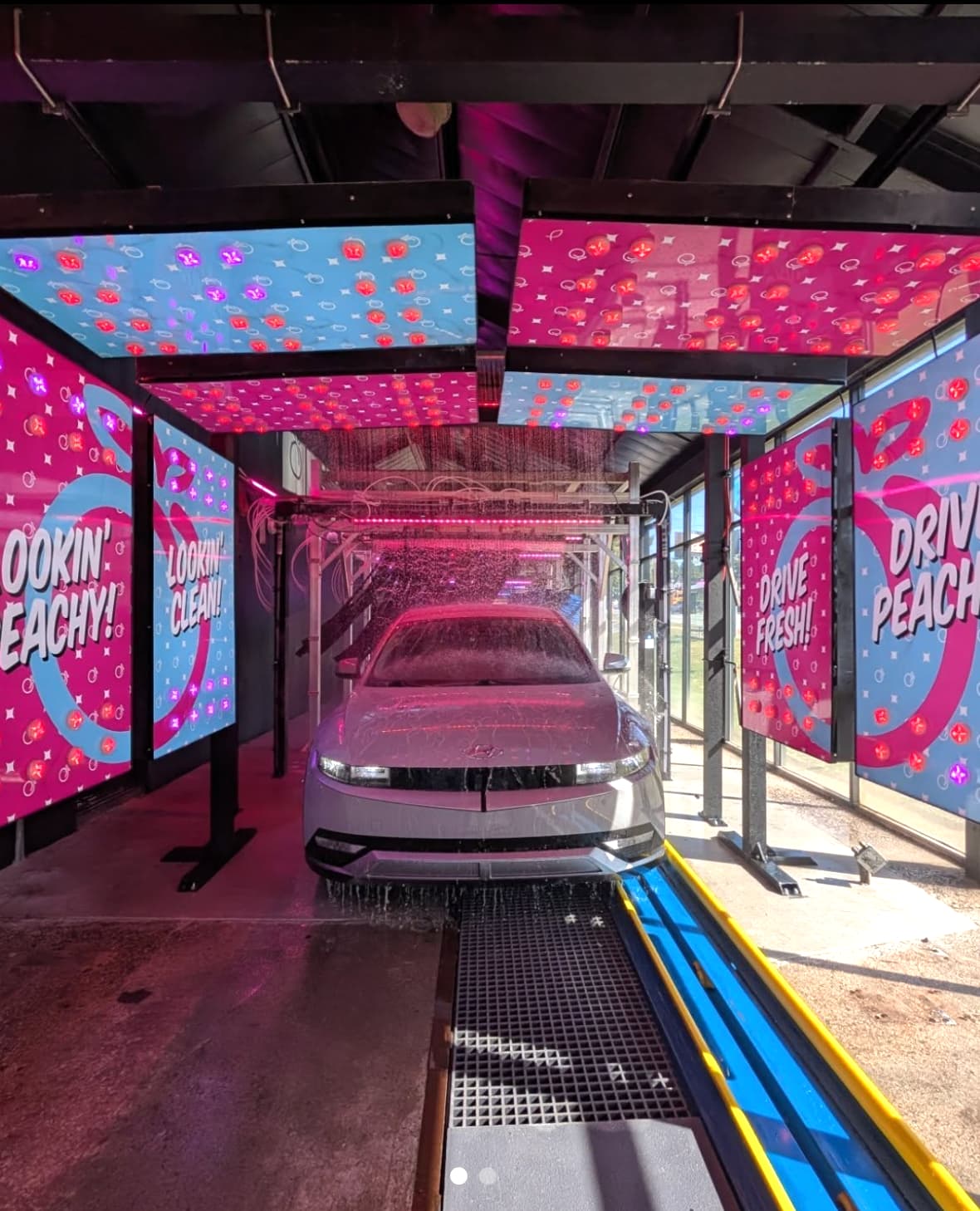 A car being washed inside the Peachy Car Wash tunnel, lit by pink and blue neon